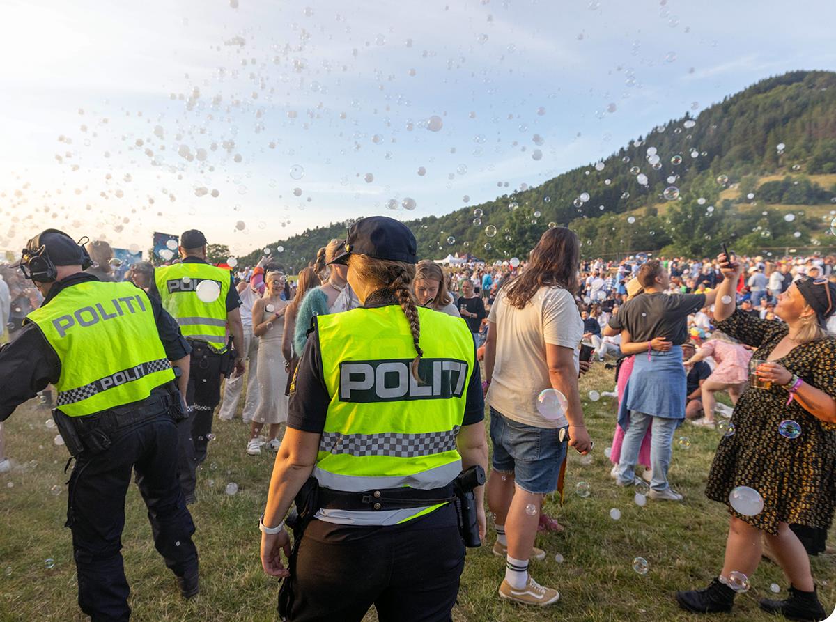 Tre politibetjenter, en kvinne og to menn, står med ryggen til kamera ute på en festival med mye folk foran en skogkledt ås. Politibetjentene har på seg caps, og gule refleksvester med teksten «Politi». Det er fint vær.