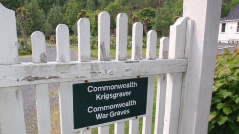 Picture of a white wooden gate at Risør war cemetery with a black sign with the following text: "Commonwealth Krigsgraver / Commonwealth War Graves".