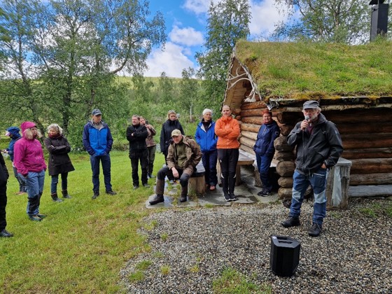 Folkeansamling utenfor laftet bygg med torvtak.