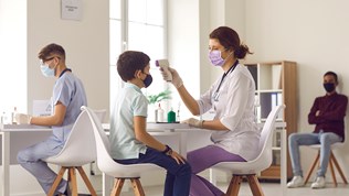 Sykepleier med munnbind holder temperaturmåleapparat opp mot en gutt