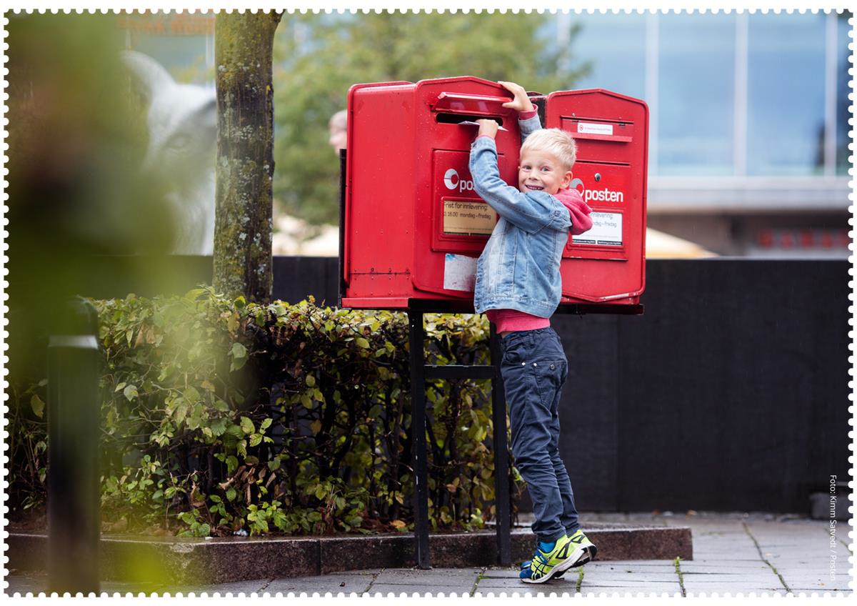 En liten gutt står på tærne og putter et brev inn i en av de to røde innleveringspostkassene fra Posten. Gutten smiler til kamera.