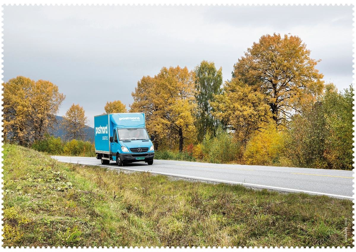 En blå PostNord-lastebil kjører bortover en vei, omgitt av trær i høstfarger. Det er overskyet, grå himmel.