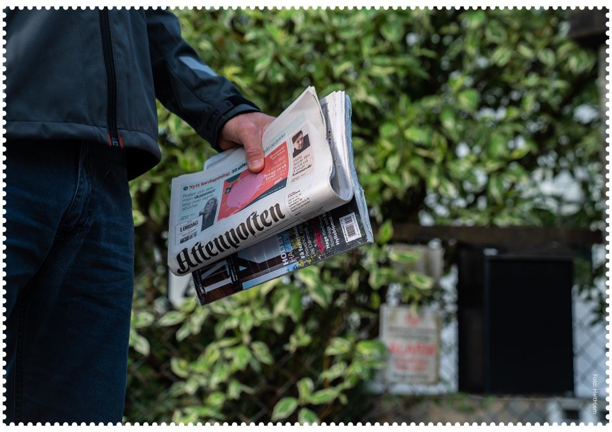 Nærbilde av en hånd som holder en Aftenposten-avis og et blad. I bakgrunnen er det en grønn hekk.Et bilde som inneholder tekst, tre, utendørs, person