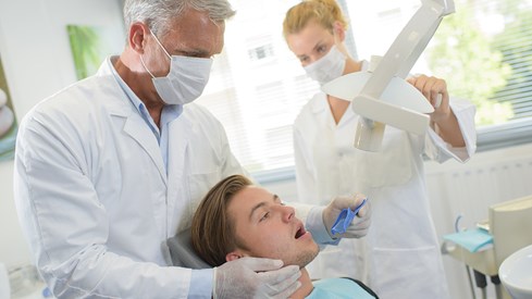 Ung mann ligger i stol på tannlegekontor, en tannlege og tannpleier står bak ham
