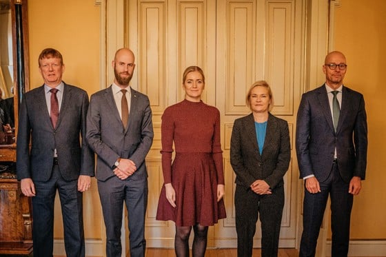 Fiver persons formally dressed standing against a yellow-tinted wall with large doors.