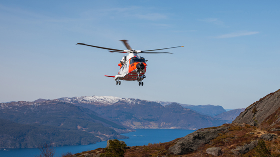 Orange and white helicopter in the air. Mountains and fjords in the background.