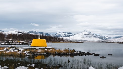 Bilde av et gult grensemerke i betong med snødekte fjell i bakgrunnen.